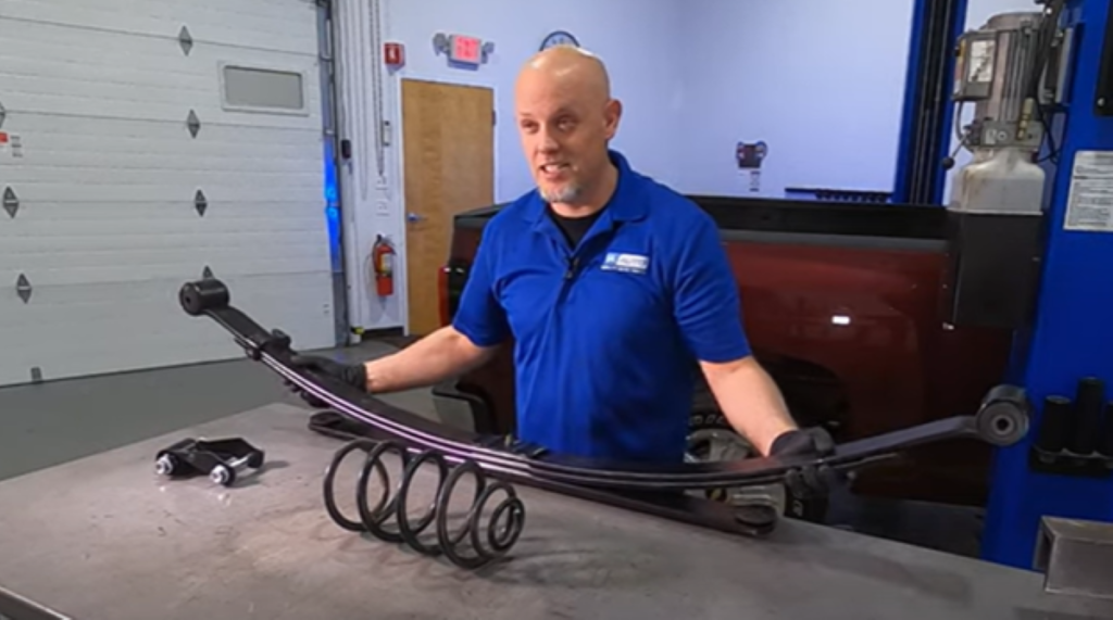 1A Auto mechanic standing before a leaf spring, shackle, and coil
